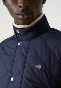 Tmavě modrá prošívaná bunda s vysokým límcem, lemovaná krémovým žebrovaným pruhem. Stříbrné patentky a vyšívané logo GANT v kontrastních barvách.