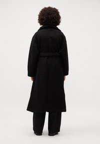 Cappotto in pile nero con vita a belted, caratterizzato da un esterno testurizzato e un ampio colletto, che arriva fino a metà polpaccio.