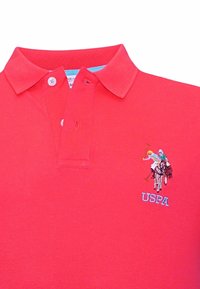 Rdeča polo majica z zloženim ovratnikom, dvema gumboma na vratu in modrim vezenim logotipom s polo igralcem na konju.