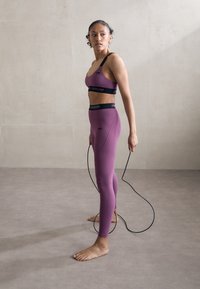 Lila sportos szett, amely sportmelltartóból és magas derekú leggingsből áll, fekete márkajelzéssel, ugrókötelet tartva, mezítláb, semleges háttér előtt.