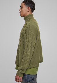 Olíva zöld kötött garbó pulóver, amely kábelkötéses mintákkal, bordás nyakkal és cropped hem-mel rendelkezik, zöld póló fölött viselve.