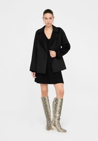 Chaqueta negra de capas con un cuello ancho, combinada con un vestido negro y botas metálicas de estampado de serpiente hasta la rodilla con punta cuadrada.