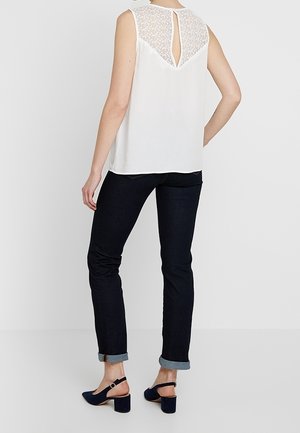 Kvinde iført hvid, ærmeløs bluse med blonde-detalje på ryggen, mørke op rullede jeans og mørkeblå slingback sko med blokhæl, stående.