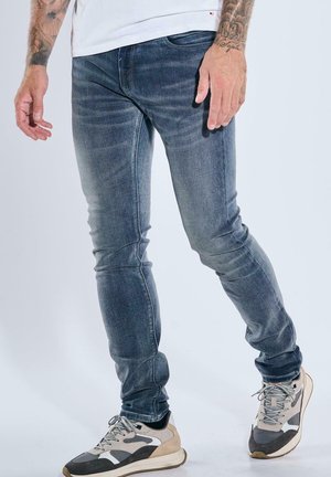 Mann trägt ausgewaschene blaue Slim-Fit-Jeans, weiße und graue Sneakers sowie ein weißes Hemd, steht mit entspannten Armen seitlich am Körper.