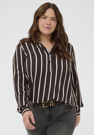 Femme aux longs cheveux bruns portant un chemisier rayé vertical noir et blanc et un jean foncé avec une ceinture noire, debout devant un fond blanc.