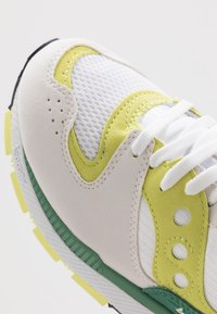 Sneaker con tomaia in mesh bianco e suede, accenti giallo brillante, dettagli verde sulla suola e lacci bianchi. Superfici testurizzate e design a strati.