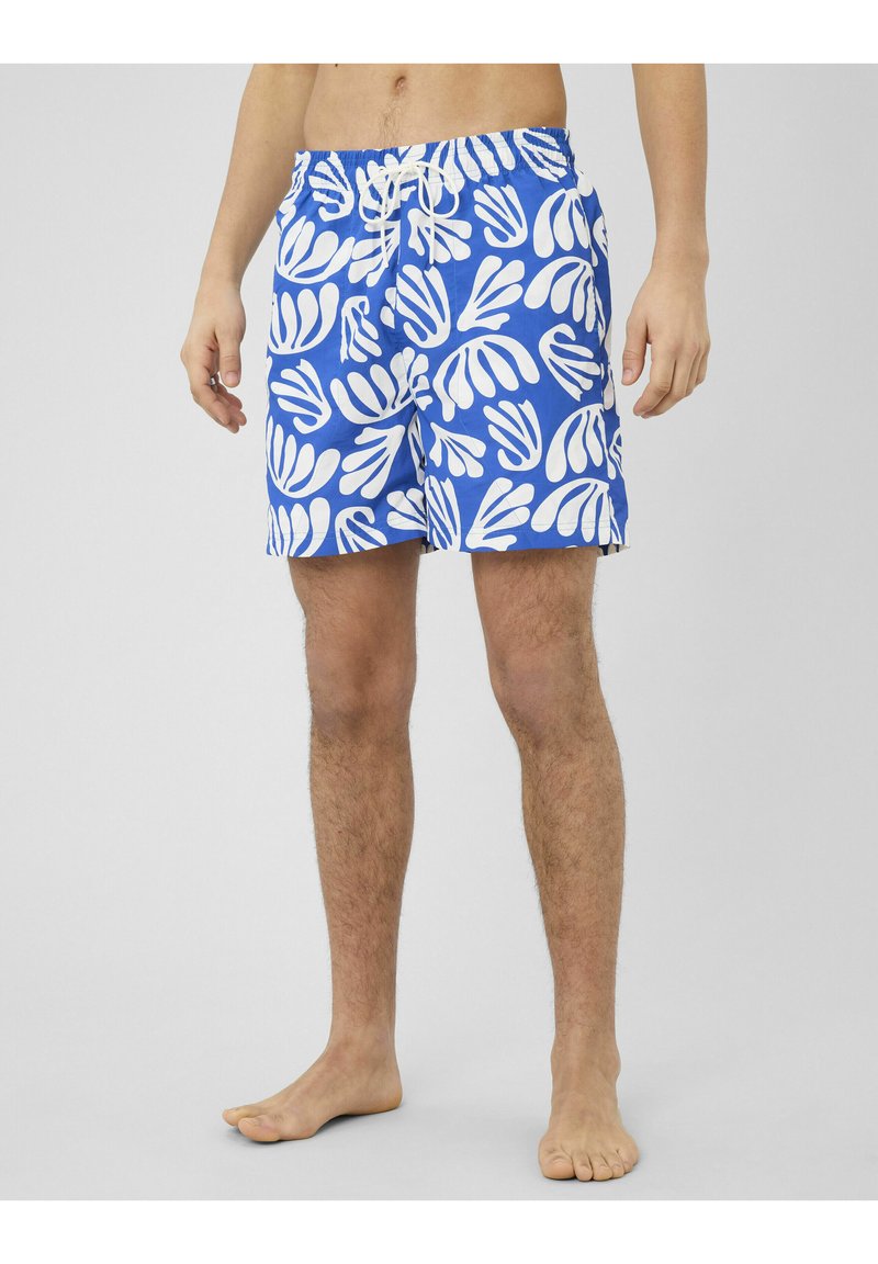 Homme portant un short de bain bleu avec des motifs abstraits de feuilles blanches, debout pieds nus sur un fond clair uni.