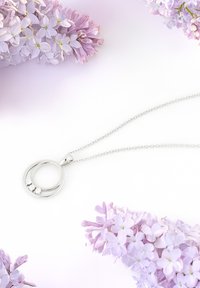Sølv halskæde med en rund vedhæng, der har tre klare ædelsten, hængende fra en delikat kæde, sat mod en lilac blomsterbaggrund.