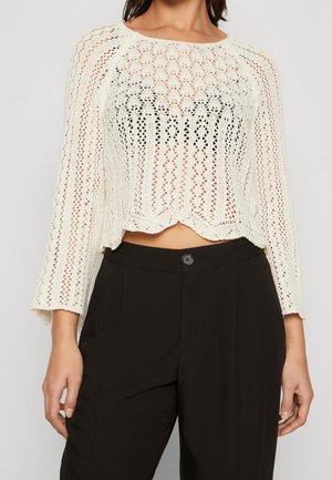 Mujer con un top corto de ganchillo blanco de manga larga combinado con pantalones negros de talle alto, de pie frente a un fondo sencillo.