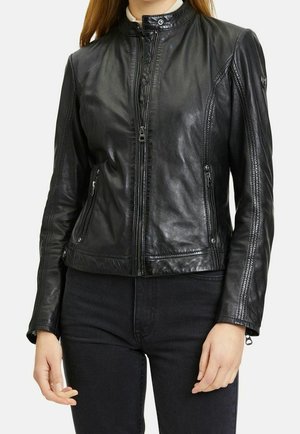 Femme portant une veste en cuir noire ajustée avec fermeture éclair devant et poches latérales, assortie à un pantalon noir sur un fond uni.