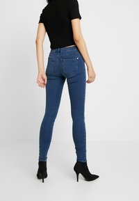 Jeans ajustados de mezclilla, con una cintura de media altura, color azul oscuro, y dos bolsillos traseros, combinados con tacones negros y una blusa negra ajustada.