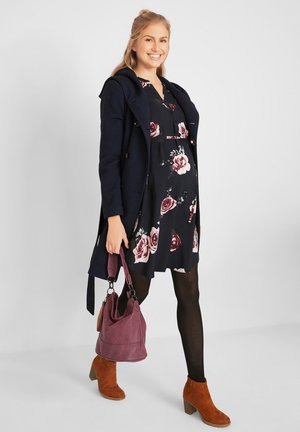 Abito floreale navy con motivi di rose, abbinato a un cappotto scuro, collant neri e stivaletti con tacco arancioni in suede. Porta una borsa viola.