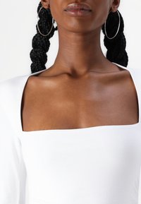 White top featuring a square neckline and fitted design. Braided hair and large hoop earrings are visible. Smooth texture.