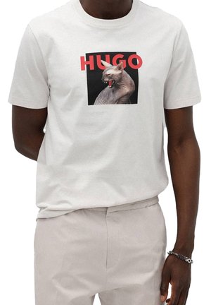 Camiseta de algodón gris claro con un gran gráfico de un gato esfinge con la palabra "HUGO" en texto rojo en negrita sobre un fondo negro.