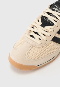 Beige tenisky s síťovaným svrškem a semišovými akcenty. Mají černé pruhy a tkaničky, spolu s hnědou gumovou podrážkou pro lepší přilnavost.