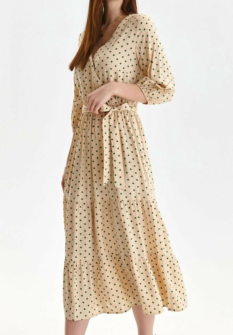 Femme portant une robe midi beige à pois verts, manches longues bouffantes, encolure en V et ceinture nouée à la taille, se tenant devant un fond blanc.