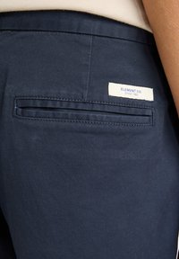 Námorné bavlnené nohavice so zadnou kapsou s horizontálnym otvorom. Štítok s nápisom "ELEMENT CO. SINCE 1992" v modrej farbe na krémovom pozadí.