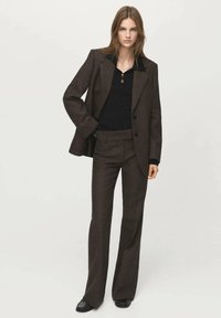 Costum maro croită, cu o jachetă structurată și pantaloni evazați, asortat cu un top negru tricotat. Prezintă reveruri mari și un detaliu cu nasturi.