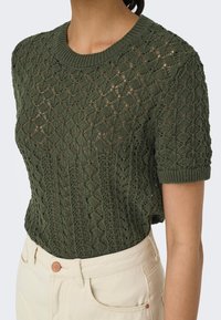 Knit short-sleeve sweater in dark green with openwork pattern, round neckline, and ribbed hem. Paired with light-colored pants.