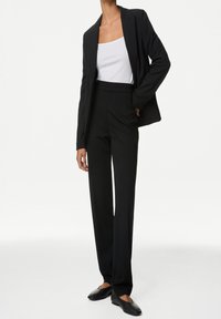 Schwarze taillierte Sakko mit Revers, kombiniert mit hoch taillierten schwarzen Hosen. Darunter ein weißes Tanktop und flache schwarze Schuhe.