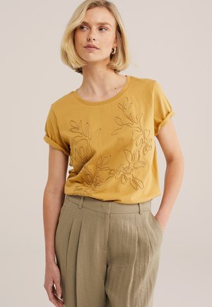 Femme aux cheveux blonds portant un t-shirt brodé de fleurs moutarde et un pantalon plissé beige, debout avec une main dans la poche.