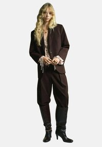 Blazer oversize marron sur un top en dentelle pâle, associé à un pantalon large marron et des bottes noires à talons hauts jusqu'aux genoux. Détails de collier complexes.
