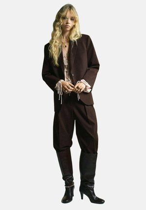 Bruine oversized blazer over een pale kanten top, gecombineerd met bruine broek met wijde pijpen en zwarte kniehoge haklaarzen. Intricate kettingdetails.