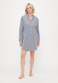 Mulher em pé descalça, vestindo uma camisa de noite de manga longa, abotoada, com riscas azuis e brancas, colarinho e bolso no peito.