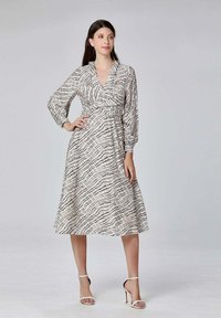 Robe à manches longues et à encolure cache-coeur en tissu crème, ornée d'un motif de lignes ondulées noires. La jupe est longue et est associée à des talons blancs.