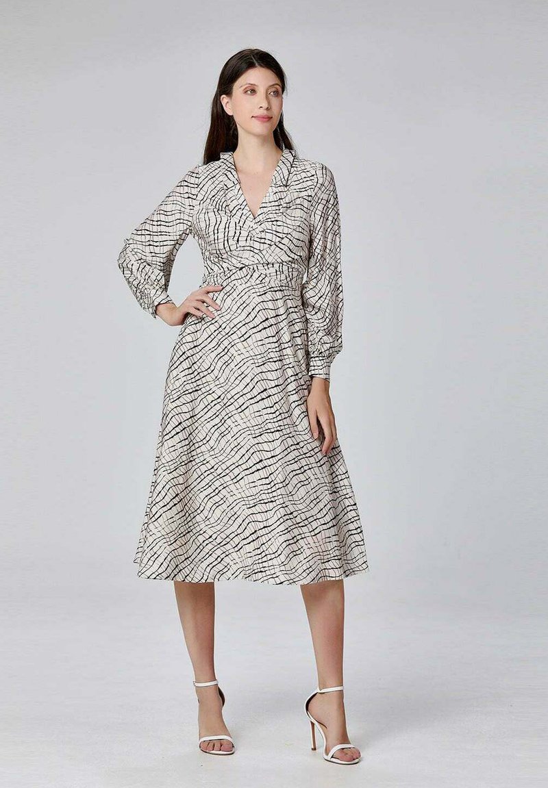 Robe à manches longues et à encolure cache-coeur en tissu crème, ornée d'un motif de lignes ondulées noires. La jupe est longue et est associée à des talons blancs.