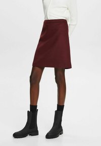 Jupe trapèze bordeaux en tissu lisse, avec une ceinture et une découpe subtile. Assortie à des bottines noires et des chaussettes.