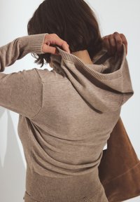 Beige stickad hoodie med ribbstickade mudd och nederkant, med huva och en mjuk textur. En brun mockaväska synlig till höger.