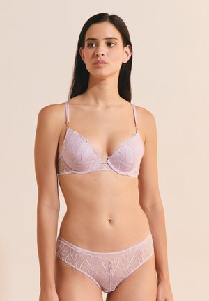Donna con lunghi capelli scuri che indossa un reggiseno e slip in pizzo rosa chiaro abbinati, in piedi davanti a uno sfondo beige uniforme.