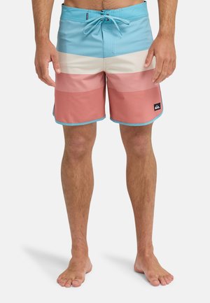 Mann trägt blau-beige-rosa gestreifte Badeshorts mit Kordelzugbund und ist barfuß vor weißem Hintergrund.