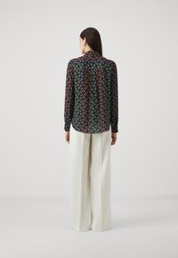 Majica s dugim rukavima u crnoj tkanini s geometrijskim uzorkom u narančastoj, zelenoj i plavoj boji, u kombinaciji sa širokim hlačama u krem boji.