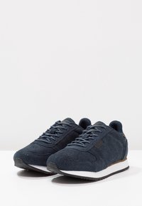 Ciemnogranatowe buty sportowe z cholewką wykonaną z teksturowanej tkaniny, białą podeszwą środkową i czarną gumową podeszwą zewnętrzną. Posiadają szare sznurowadła i subtelny akcent na pięcie.