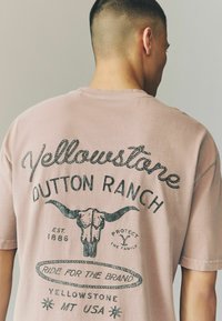 Lyserød bomulds t-shirt med et vintage design, der har sort trykt tekst, herunder "Yellowstone Dutton Ranch" og et grafik af en oksehoved.