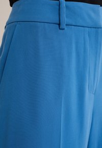 Modré látkové kalhoty s hladkým povrchem, s plochým pasem a boční kapsou, vystavené pod úhlem pro zdůraznění střihu a barvy.