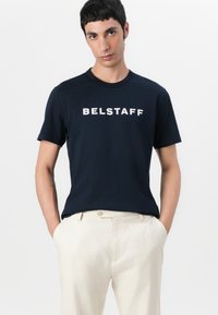 Kļavi zils kokvilnas T-krekls ar baltu "BELSTAFF" logo, apaļu kakla izgriezumu, īsām piedurknēm, valkāts ar gaiši bežām biksēm. Gluda tekstūra.