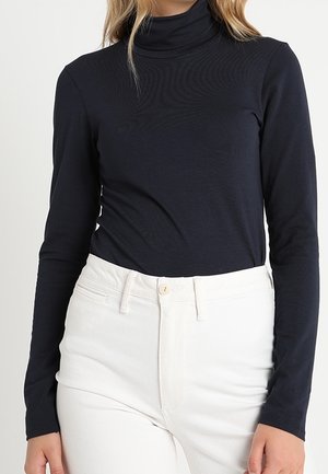 Woman wearing a fitted black long-sleeve turtleneck tucked into high-waisted white pants with a button and zipper closure.