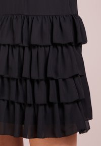 Czarna, wielowarstwowa sukienka z kilkoma falbanami wykonana z lekkiego materiału; posiada przezroczysty dół i starannie wykończone krawędzie dla podkreślenia detali.