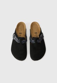 Black suede clogs with a round toe, featuring a braided strap with a metal buckle. The insoles are textured and tan in color.