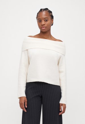 Suéter crema de hombros caídos con cuello acanalado, hecho de una tela suave, presenta un corte relajado y una textura sutil. Combinado con pantalones de rayas.