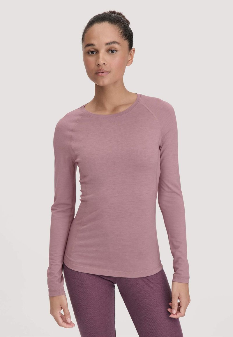 Woman wearing dusty pink long-sleeve fitted top and textured purple leggings, standing against a plain light background.