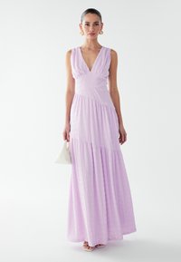Lavender maxi dress with a V-neck, textured fabric, and gathered bodice; flared skirt and subtle tiered design; paired with a white bag.
