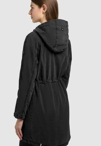 Femme vue de dos portant une longue veste noire à capuche avec des détails de coutures apparentes et une taille élastique.