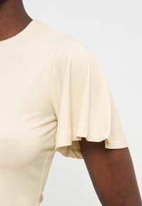 Μπλούζα σε εκρού χρώμα με κοντά μανίκια και λεπτομέρεια βολάν, φορεμένη στον άνω βραχίονα και τον ώμο ενός ατόμου με σκουρόχρωμο δέρμα, πάνω σε απλό φόντο.