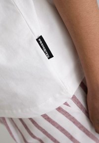 White cotton shirt with a curved hem, featuring a black "PROHIBITED" label on the side, paired with red-striped fabric below.