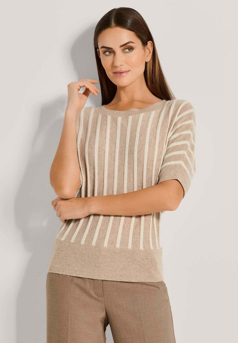 Vrouw met bruin haar die een beige sweater met korte mouwen en witte verticale strepen draagt, samen met beige broek, staand tegen een lichte muur.