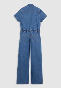 Vue arrière d'une combinaison en denim bleu à manches courtes, jambes larges, taille élastique, deux poches arrière et col sur fond gris clair.